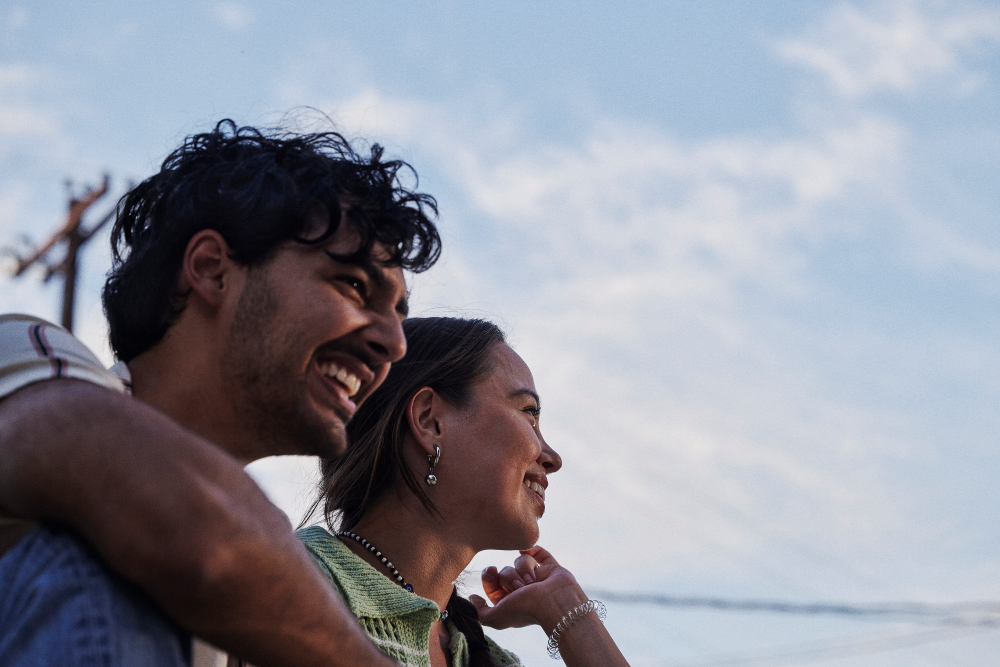 Two people smile and look off into the distance under a blue sky with soft clouds. One person has an arm around the other's shoulder, and both appear happy and relaxed outdoors.