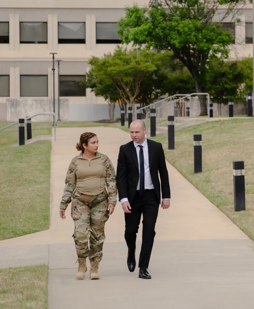 A woman in camouflage uniform walks next to a man in a black suit, on a sidewalk in front of a building.