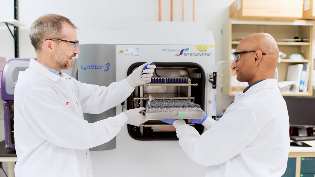 Two scientists in white lab coats and gloves work together. One holds a tray of samples while the other uses a pipette, with laboratory equipment and shelves visible in the background.