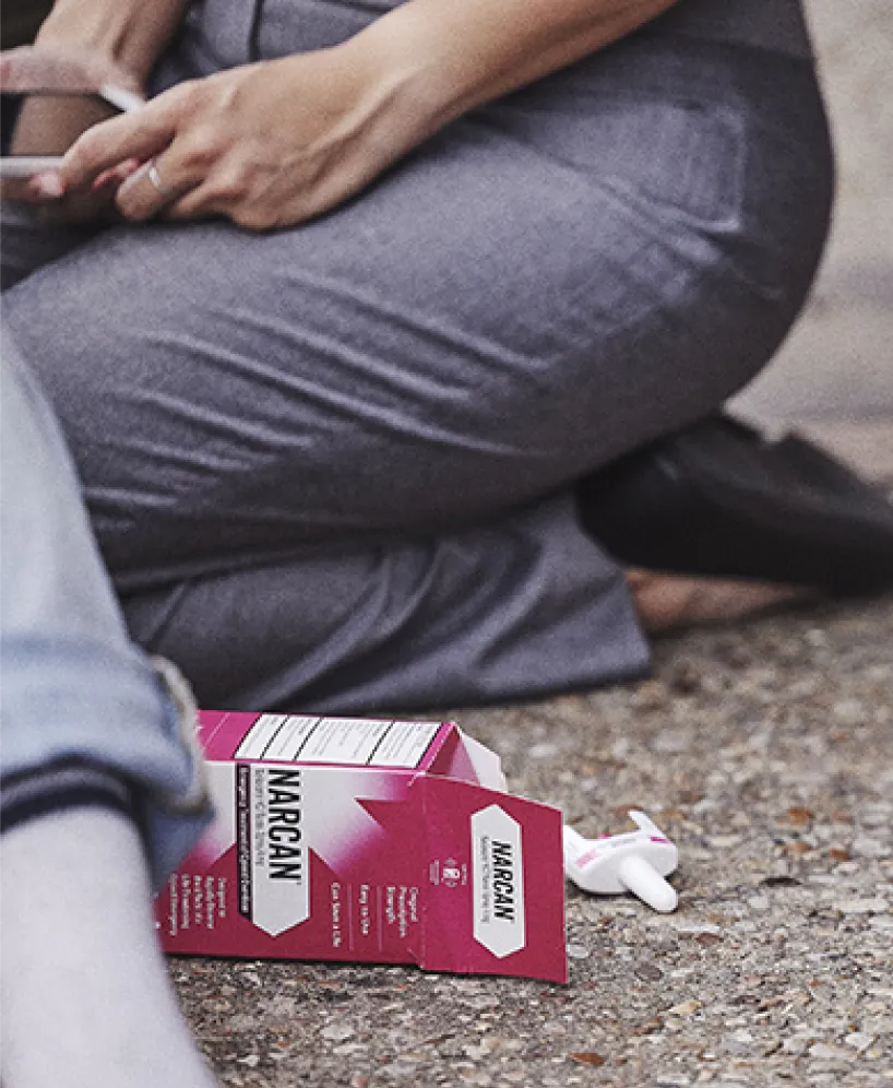 A person kneels on the ground next to an open Narcan (naloxone) nasal spray box with a nasal applicator visible, suggesting a medical emergency or overdose intervention scene.
