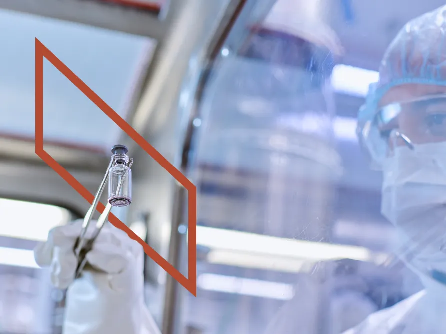 A scientist in protective gear holds a vial with metal tongs inside a laboratory, viewed through a transparent glass screen with a red outlined square.