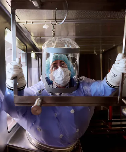 A person wearing protective clothing, gloves, a hair net, and a face mask works inside a sterile laboratory environment, handling equipment through a sealed glass chamber.