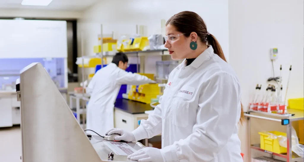 A scientist wearing a white lab coat, gloves, and safety glasses operates equipment in a laboratory. Another person works at a bench in the background. The lab is bright and filled with yellow storage bins.
