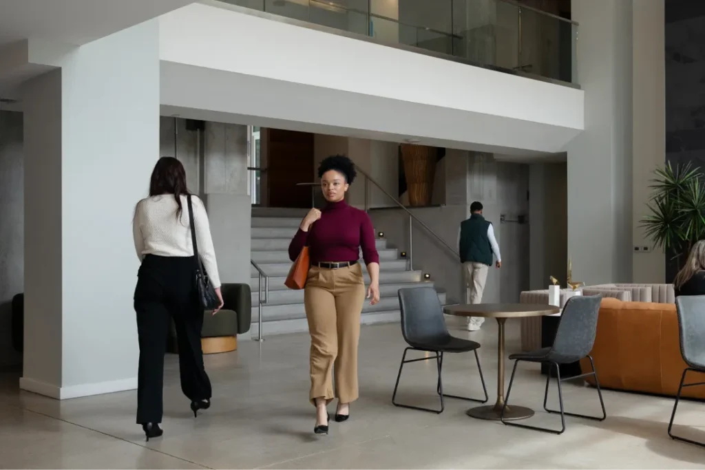 A woman in a burgundy top and tan pants walks through a modern lobby with a staircase, seating area, and people in the background.