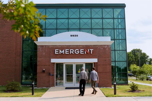 Two people walk toward a brick building with a glass facade and the word “EMERGENT” above the entrance. Trees and greenery are visible around the building.
