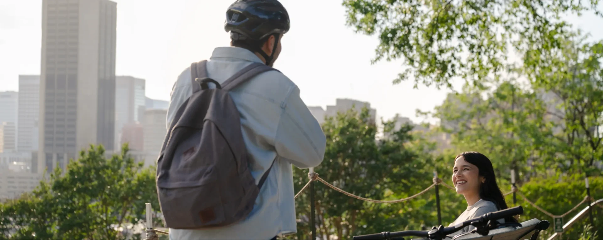 A person wearing a helmet and backpack stands with a bicycle, talking to a smiling woman sitting on a park bench, with city buildings and green trees in the background.