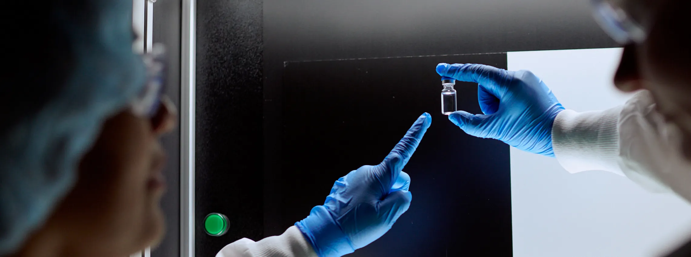 Two people wearing blue gloves and lab coats examine a small glass vial in a laboratory setting, with one person holding the vial up to the light and the other pointing at it.