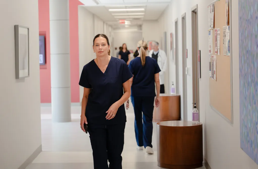 A woman in navy scrubs walks down a brightly lit hospital hallway. Other medical staff in similar uniforms and a doctor are visible in the background.