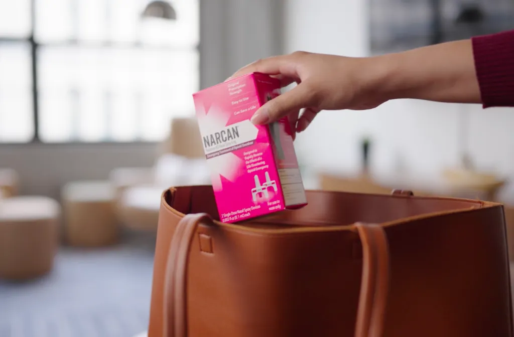 A hand places a pink Narcan nasal spray box into a brown leather bag, with a blurred indoor background.