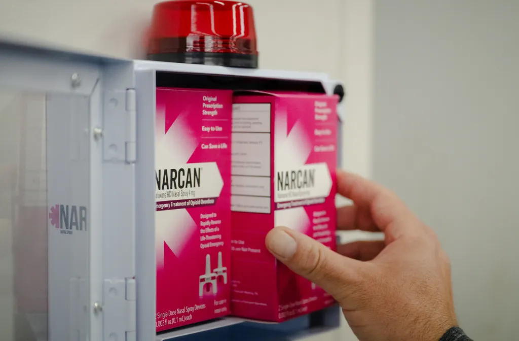 A hand reaches into a wall-mounted box containing Narcan nasal spray kits, with a red warning light on top of the box.