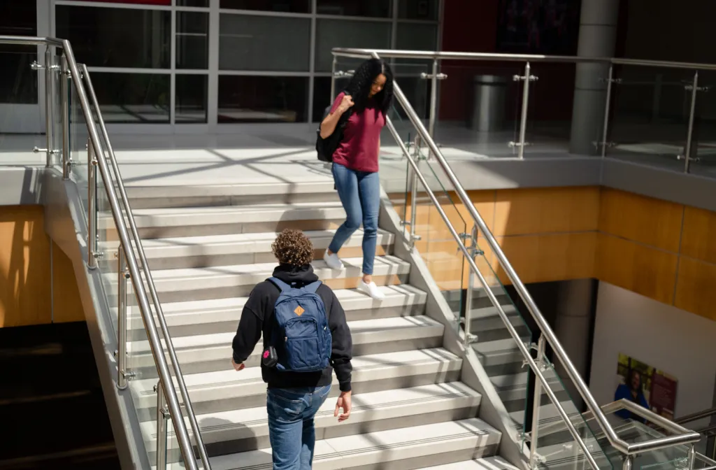Two students walk on a staircase inside a modern building; one is going up with a backpack, while the other is coming down, also carrying a backpack. Sunlight streams through large windows.