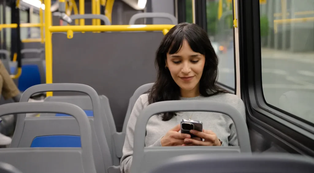 A woman with long dark hair sits alone on a bus, smiling while looking at her smartphone. She is wearing a light gray sweater and is seated next to the window. The bus appears mostly empty.
