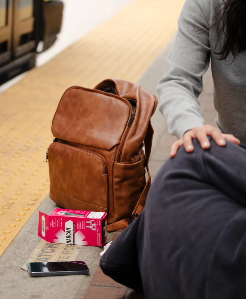 A person sits on a train platform beside a brown backpack, a juice box, a phone, a ticket, and a dark blanket, with a train visible in the background.
