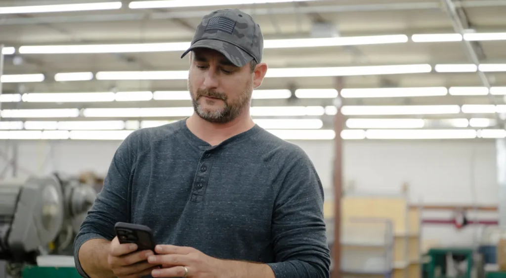 Man in a gray cap and long-sleeve shirt standing in an industrial factory setting, looking down at a smartphone he is holding with both hands.