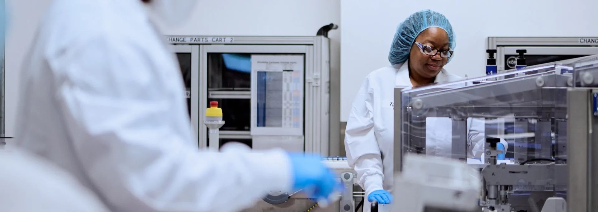 Two scientists in protective clothing and hairnets work in a laboratory with scientific equipment and machines. One scientist is in focus, looking at a device, while the other is blurred in the foreground.
