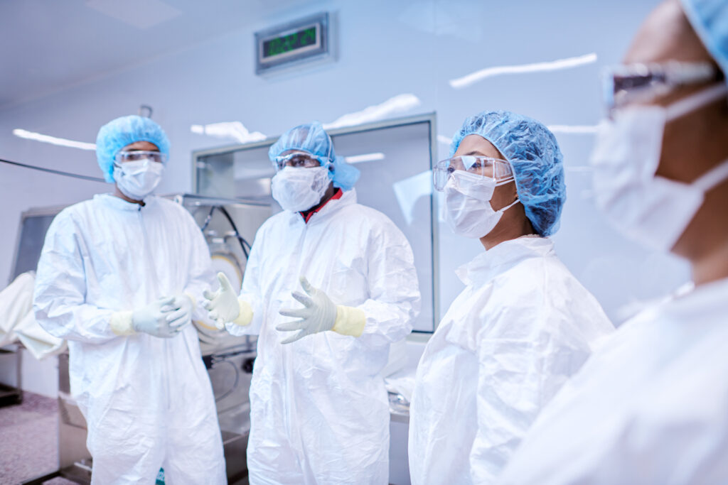 Four people in protective suits, gloves, masks, and goggles stand in a lab, having a discussion. Laboratory equipment and a clock are visible in the background.